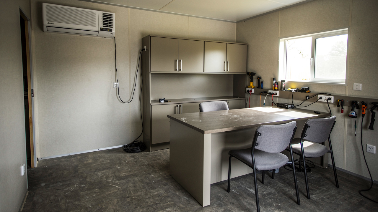 Intérieur d’un bungalow bureau aménagé sur un chantier, avec climatisation, bureau, chaises simples et rangements, éclairage naturel et mobilier pratique.